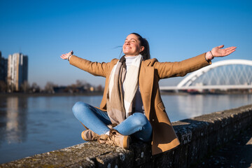 Beautiful woman in warm clothing with her arms outstretched  enjoys resting by the river on a sunny winter day.