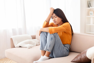 Upset young asian woman sitting on couch, touching her head - obrazy, fototapety, plakaty