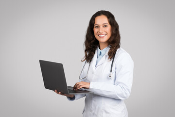 Online consultation service. Confident woman doctor using laptop, standing on grey studio...