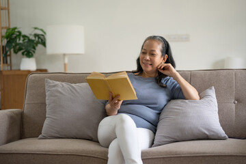 Senior asian old woman sitting on sofa in the living room reading a book to relax on a leisure day.