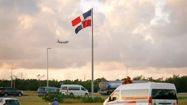 The national flag of the Dominican Republic waving in the wind on the roadside and landing airplane in the sunset sky. Many cars on Punta Cana highway in peak season of summer tourist vacations 