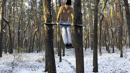 Strong and muscular man doing exercises on horizontal bar at winter forest. Young sportsman...
