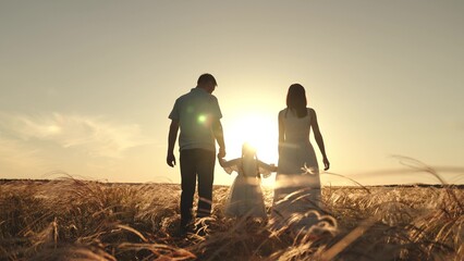 Silhouettes of spouses with daughter walk along field of wheat in evening enjoying time in circle...