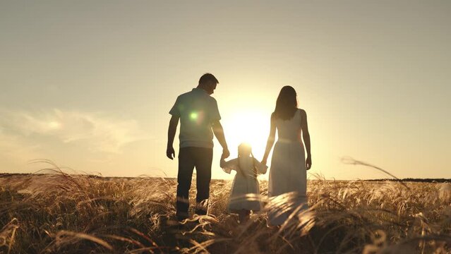 Silhouettes of spouses with daughter walk along field of wheat in evening enjoying time in circle of family. Silhouettes of father and mother with girl in light summer dress with long braids at sunset