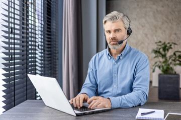 Professional middle-aged man with headset working on laptop in a modern office setting, showcasing...