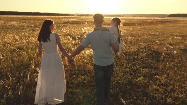 Daughter sitting in arms of father walking with wife through summer wheat field at sunset. Daughter with braids sitting in arms of father enjoying company of happy family and summer sunshine