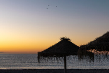 Sunset on the beach with thatched umbrellas and seagulls. Nerja, Costa del Sol, Spain.