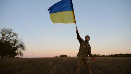 Young male military in uniform jogging with flag of Ukraine on meadow at dusk. Soldier of ukrainian army waving blue-yellow banner in honor of victory against russian aggression. 