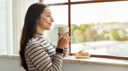 Contented young woman enjoying a peaceful moment, sipping coffee and gazing out a sunny window