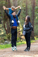 Fototapeta premium Happy family walking in the forest. The father jumps up with his son on his shoulders, the child laughs with joy, and the mother looks on as the father plays with his son
