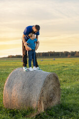 Portrait of a father and son standing on a haystack and looking at each other in a rural field in...