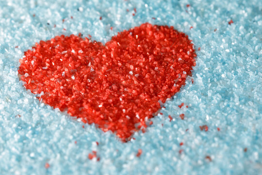 The shape of a red heart on a light blue background is made of colored sand crystals. Photo for background, with small depth of field