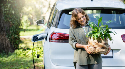 Woman with plant in pot next to a charging electric car in the yard of a country house