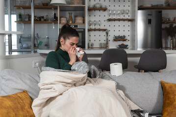Young sick woman wrapped in a blanket wipes her nose with paper tissues, got sick because she...