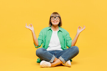 Full body happy elderly woman 50s years old wear green shirt glasses casual clothes sitting hold spread hands in yoga om aum gesture relax meditate try calm down isolated on plain yellow background.