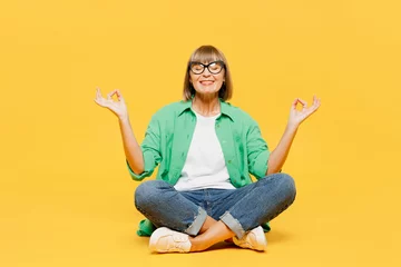  Full body happy elderly woman 50s years old wear green shirt glasses casual clothes sitting hold spread hands in yoga om aum gesture relax meditate try calm down isolated on plain yellow background. © ViDi Studio