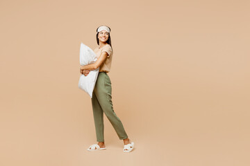 Fototapeta premium Full body side view young calm Latin woman she wears pyjamas jam sleep eye mask rest relax at home hold pillow look aside on area isolated on plain pastel beige background Good mood night nap concept