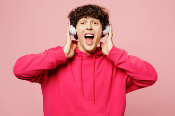 Young surprised shocked happy Caucasian man he wears hoody casual clothes listen to music in headphones look camera isolated on plain pastel light pink background studio portrait. Lifestyle concept.