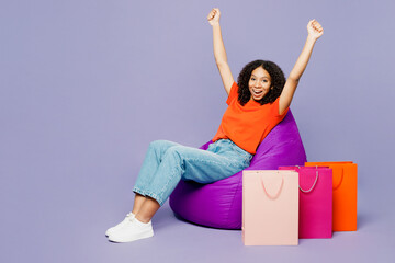 Winner little kid teen girl of African American ethnicity wear orange t-shirt sit in bag chair hold shopping paper package bags isolated on plain purple background. Black Friday sale buy day concept.