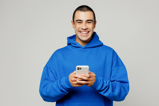 Young smiling happy middle eastern man he wearing blue hoody casual clothes hold in hand use mobile cell phone look camera isolated on plain solid white background studio portrait. Lifestyle concept.