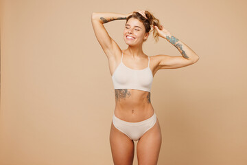 Young calm nice lady woman with slim body perfect skin wear nude top bra lingerie stand looking aside holding hair in hands isolated on plain pastel light beige background. Lifestyle diet fit concept.
