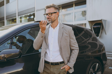 Side view adult man customer male buyer client wearing white shirt classic grey suit stand near black electric car while charging record voice message on mobile cell phone outside. Business concept.