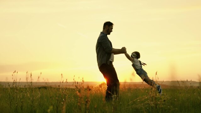 Happy family, dad plays with child as pilot, circling child by hands, kid rejoices and laughs. Silhouette of father and child, little girls, playing, enjoying sunset in park in nature on summer day.