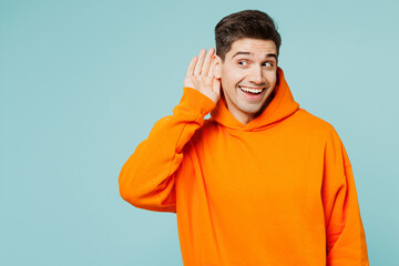 Young curious nosy man he wearing orange hoody casual clothes try to hear you overhear listening intently isolated on plain pastel light blue cyan color background studio portrait. Lifestyle concept.