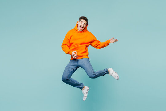 Full body happy singer young man he wears orange hoody casual clothes jump high play do air guitar gesture isolated on plain pastel light blue cyan color background studio portrait. Lifestyle concept.