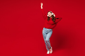 Full body young woman she wearing knitted sweater white hat casual clothes listen to music in headphones dance raise up hand isolated on plain red color background studio portrait. Lifestyle concept. - obrazy, fototapety, plakaty
