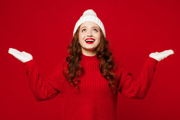Young happy smiling fun woman wears knitted sweater white hat gloves casual clothes look overhead spread hands pov catch snow flakes isolated on plain red color background studio. Lifestyle concept.