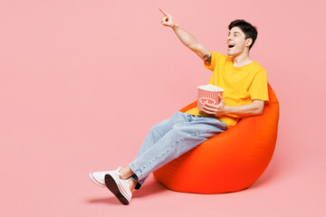Full body young man wear yellow t-shirt casual clothes sit in bag chair watch movie film eat popcorn point index finger aside isolated on plain pastel light pink background studio. Lifestyle concept