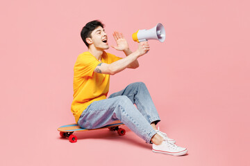 Full body young man wear yellow t-shirt casual clothes sit on skateboard scream in megaphone announces discounts sale Hurry up isolated on plain pastel light pink background studio. Lifestyle concept
