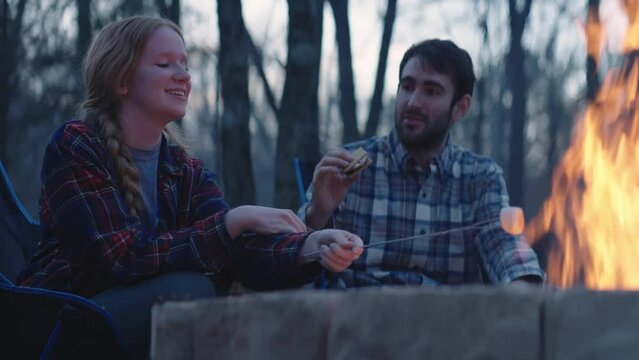 Young man and woman sit beside a fire and make s&rsquo;mores 