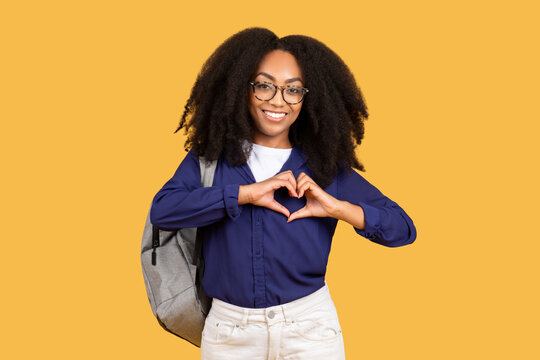 Happy black lady student with backpack, making heart shape with hands on chest, on yellow background