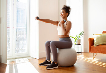 Sporty millennial lady with dumbbells exercising on stability ball indoor
