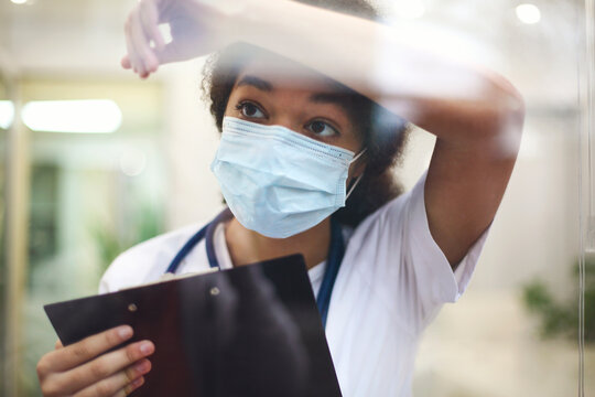 Young tired overworked african american medical worker in protective face mask looking out of window