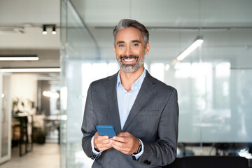 Smiling mature Latin businessman holding smartphone in office looking at camera. Middle aged manager using cell phone mobile app. Digital technology application and solutions for business. Copy space