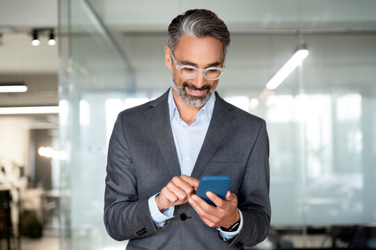 Close-up of smiling mature Latin or Indian businessman holding smartphone in office. Middle aged manager using cell phone mobile app. Digital technology application and solutions for business concept.