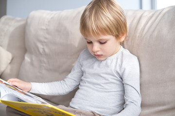 little boy reading book, sitting on sofa at home