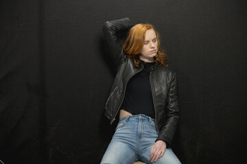 Young woman with red hair in black jacket and jeans poses in studio before a black background