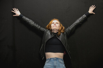Young woman with red hair in black jacket and jeans poses in studio before a black background