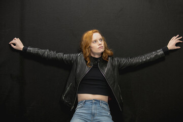 Young woman with red hair in black jacket and jeans poses in studio before a black background