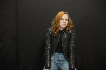Young woman with red hair in black jacket and jeans poses in studio before a black background