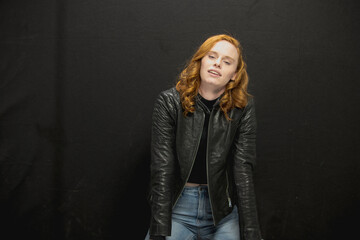Young woman with red hair in black jacket and jeans poses in studio before a black background