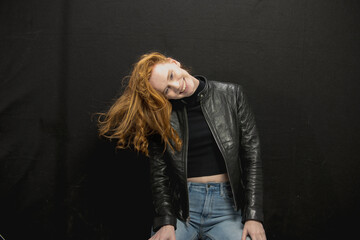 Young woman with red hair in black jacket and jeans poses in studio before a black background