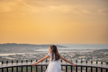 Girl in a first communion dress celebrating her day on a viewpoint with the city and the bay in the background during sunset