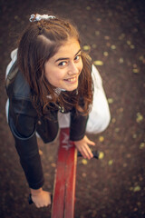 Fototapeta premium Little girl in communion dress and black leather jacket playing on a swing in a public park