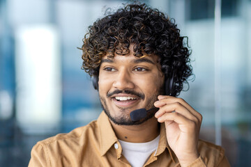 A happy male customer service agent with curly hair smiling, wearing a headset in an office environment.