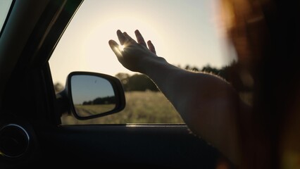 Girl with long hair is sitting in front seat of car, stretching her arm out window and catching...